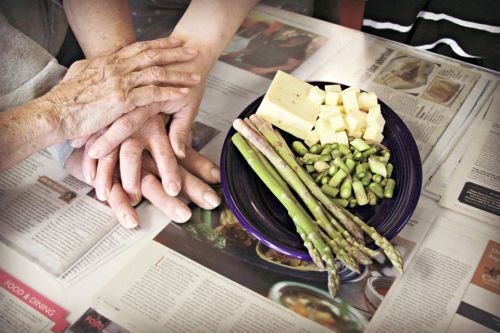 Two hands resting on a table with a plate of asparagus and cheese cubes, surrounded by newspapers.