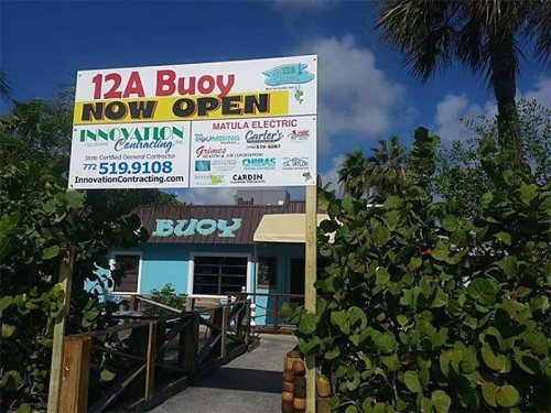 Sign for "12A Buoy" with "NOW OPEN" and various business logos, surrounded by greenery and a blue sky.