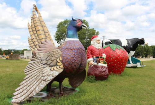 Colorful oversized sculptures of a peacock, Santa Claus, and a strawberry in a grassy field under a cloudy sky.