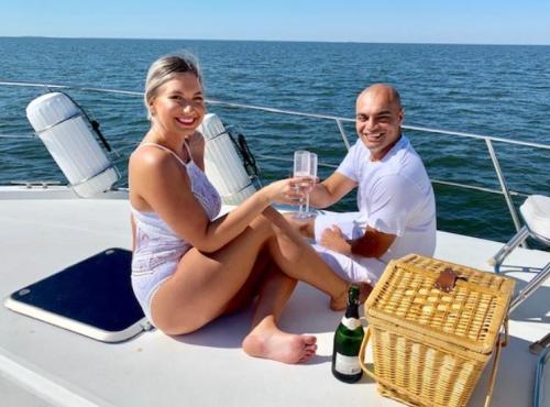 A couple enjoys a sunny day on a boat, smiling and toasting with drinks, with a picnic basket nearby.