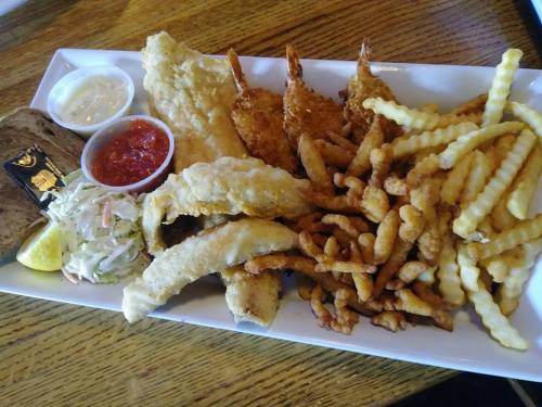 A platter of fried fish, shrimp, crinkle-cut fries, coleslaw, and dipping sauces, served with a slice of bread.