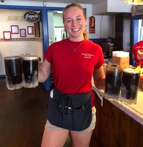 A smiling waitress in a red shirt holds four large glasses of soda at a restaurant.