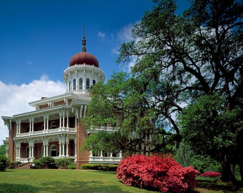 Historic building with a red dome, surrounded by lush greenery and vibrant pink flowers under a blue sky.