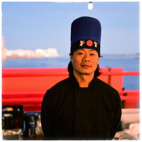 A chef wearing a blue hat and black uniform stands against a scenic waterfront backdrop.