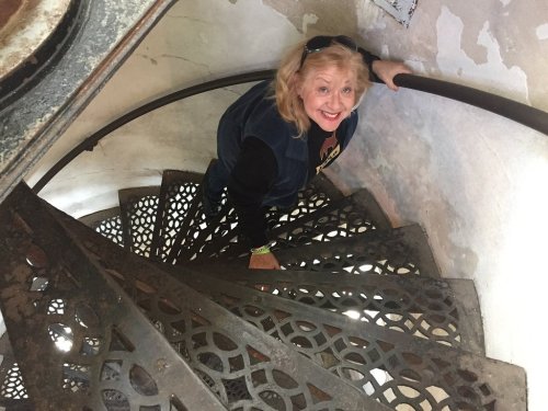 A smiling woman climbs a spiral staircase with decorative metal steps, surrounded by a weathered wall.