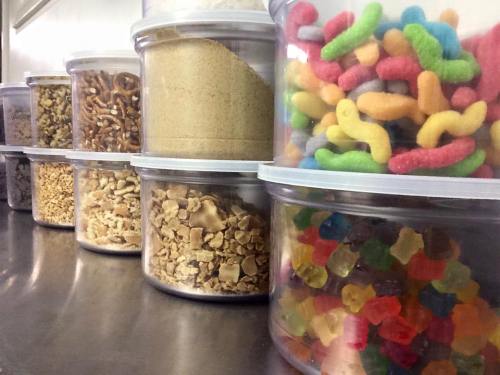 Containers filled with various snacks and candies, including gummies, pretzels, and granola, lined up on a table.