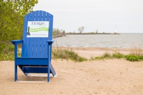 A large blue adirondack chair on a sandy beach with a sign for Huron Beach and a distant lighthouse by the water.