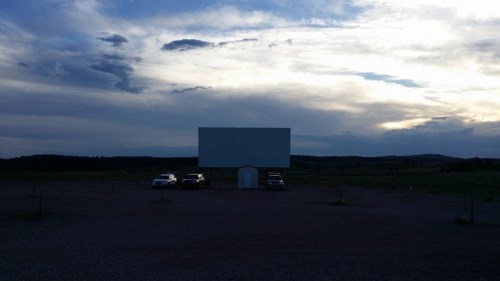 A blank outdoor movie screen at dusk, with a few cars parked nearby against a cloudy sky.