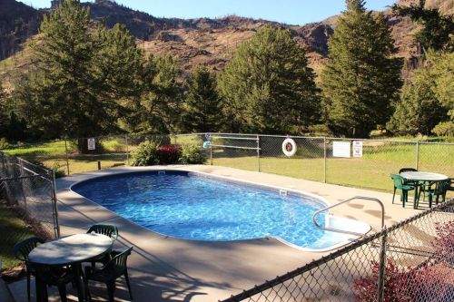 A clear swimming pool surrounded by a fence, with green trees and mountains in the background. Tables are nearby.