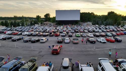 Aerial view of a busy drive-in movie theater with numerous cars parked and people gathering outside as the sun sets.