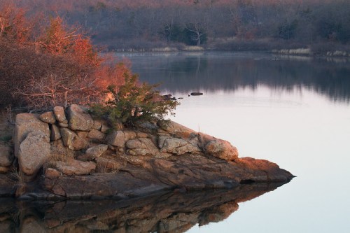 A rocky shoreline reflects in calm water, surrounded by trees and a serene landscape at sunset.