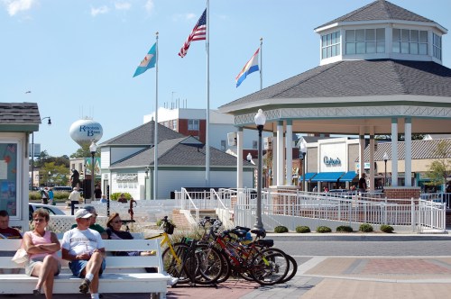 A sunny boardwalk scene with people sitting on benches, bicycles parked, and flags flying near a gazebo.