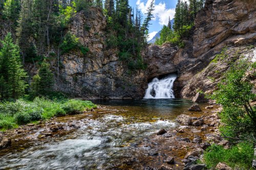 A serene waterfall cascades into a clear pool, surrounded by rocky cliffs and lush greenery under a bright blue sky.
