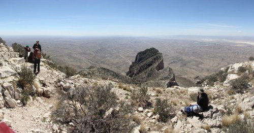 A panoramic view of a mountainous landscape with hikers on rocky terrain and a distant peak under a clear blue sky.