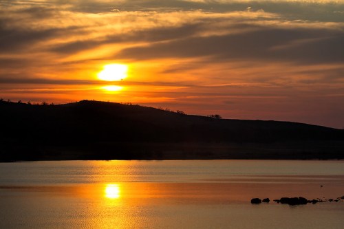 Sunset over a calm lake, with vibrant orange and yellow hues reflecting on the water and silhouetted hills in the background.