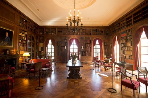 A spacious library with wooden shelves filled with books, elegant furniture, and large windows letting in natural light.
