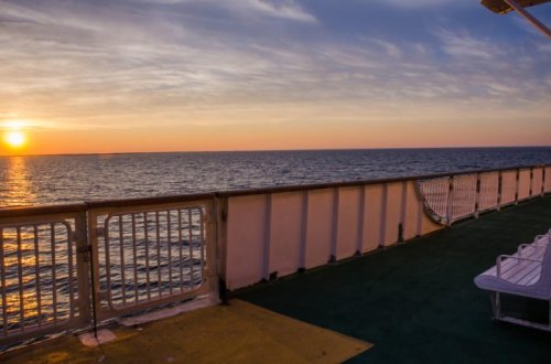 Sunset over calm waters viewed from a ship's deck, with a railing and empty seating in the foreground.