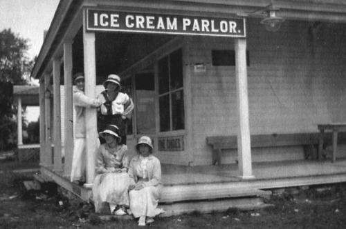 Black and white photo of an ice cream parlor with four people, two seated and two standing, all in early 20th-century attire.
