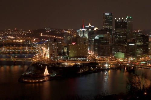 Night view of Pittsburgh skyline with illuminated buildings and a decorated tree by the river.