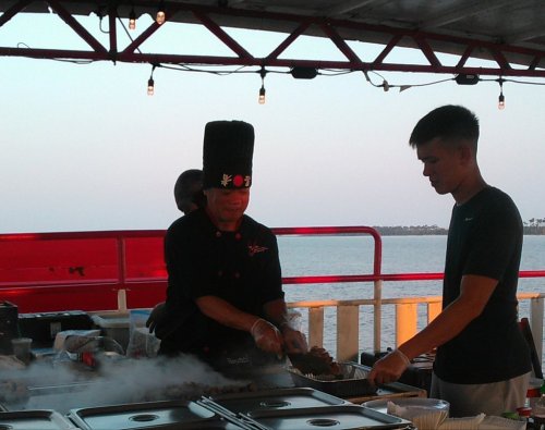 A chef in a tall hat cooks on a grill while a young man watches, with a scenic waterfront in the background.