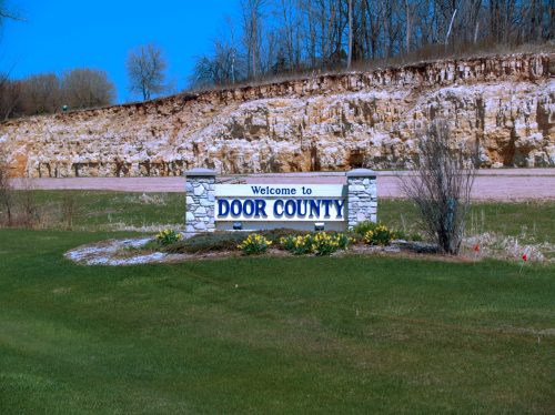 A welcome sign for Door County, surrounded by green grass and a rocky hillside under a clear blue sky.