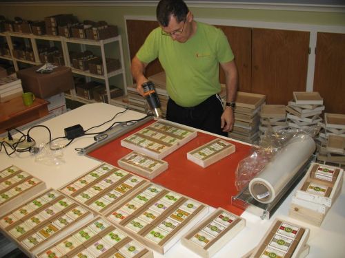A man in a green shirt uses a heat gun on wooden boxes arranged on a table in a workshop.