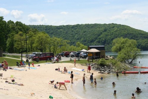 A sandy beach with people swimming and playing, surrounded by green hills and parked cars in the background.
