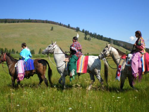 Three individuals on horseback, dressed in colorful traditional attire, riding through a green, grassy landscape.