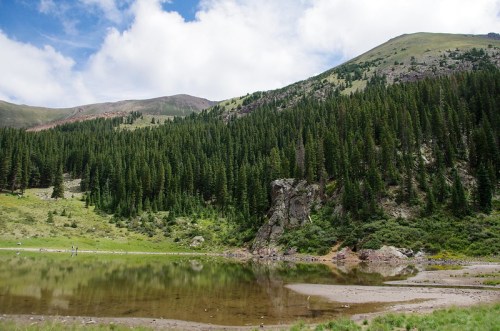 A serene landscape featuring a calm lake surrounded by lush green forests and mountains under a partly cloudy sky.