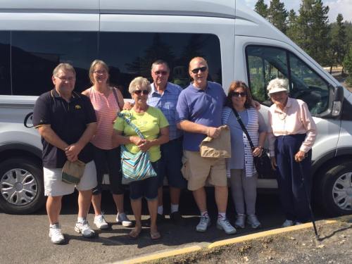 A group of seven people stands beside a van, smiling and posing for the photo in a sunny outdoor setting.