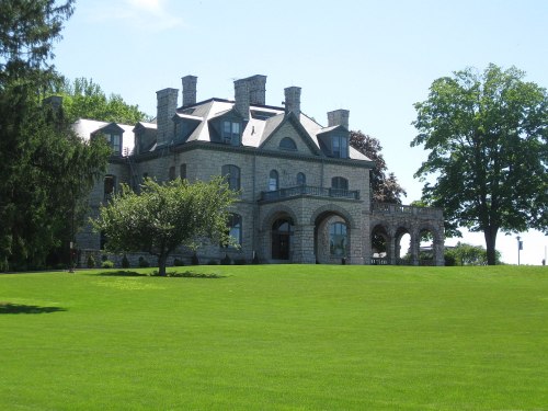 A large stone mansion with multiple chimneys, surrounded by lush green grass and trees under a clear blue sky.