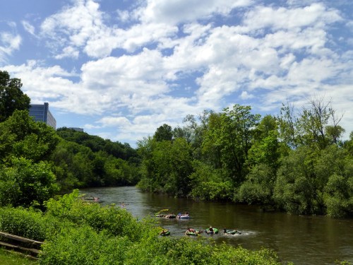 A scenic river surrounded by lush greenery and trees, with people kayaking under a bright blue sky and fluffy clouds.