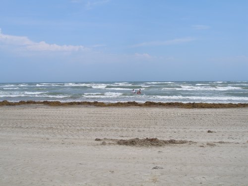 A sandy beach with gentle waves and two people wading in the water, under a clear blue sky.