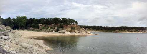 A panoramic view of a calm lake surrounded by rocky shores and lush greenery under a cloudy sky.