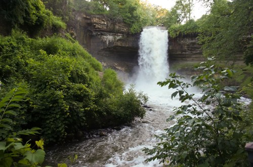 A waterfall cascades over a rocky ledge, surrounded by lush greenery and flowing water below.