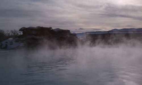 A serene hot spring surrounded by mist, with rocky formations and a cloudy sky in the background.