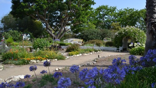 A vibrant garden with colorful flowers, a winding path, and a white bench under a large tree, surrounded by greenery.