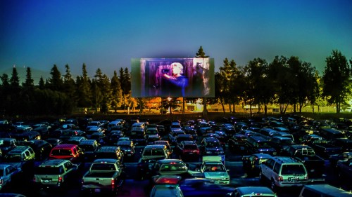 A drive-in movie theater at dusk, with a large screen showing a film and cars parked in front.