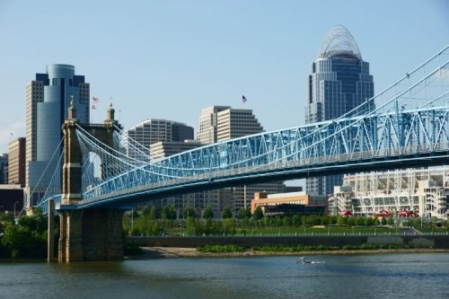 A blue suspension bridge spans a river with a city skyline and modern buildings in the background.