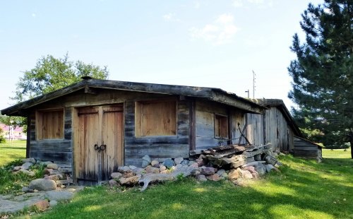 A rustic wooden cabin with boarded windows, surrounded by grass and rocks, under a clear blue sky.