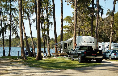 A green truck parked near an RV by a lake, surrounded by tall trees and a grassy area.