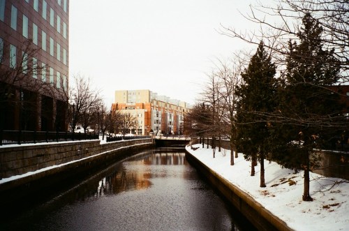 A snowy canal lined with trees and buildings, reflecting the winter landscape under a cloudy sky.