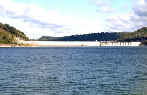 A view of a dam across a lake, surrounded by hills and a partly cloudy sky.