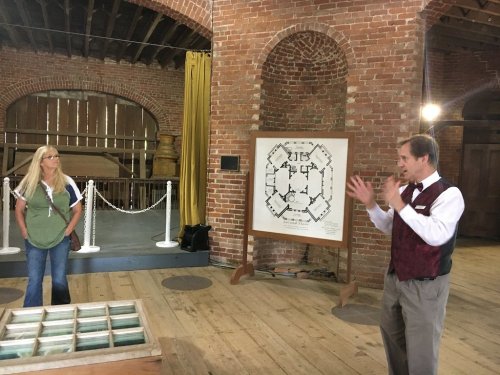 A man in a vest gestures while speaking to a woman in an apron, with a historical building and a diagram in the background.