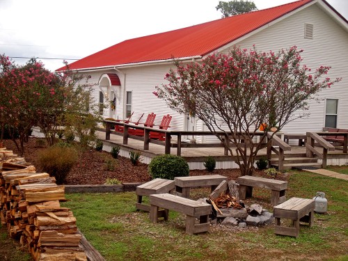 A cozy building with a red roof, surrounded by flowering bushes and a wooden fire pit area with benches.