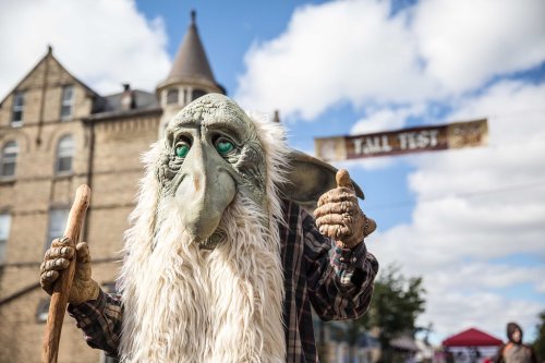 A whimsical creature in a mask and costume stands outdoors, giving a thumbs up with a historic building in the background.