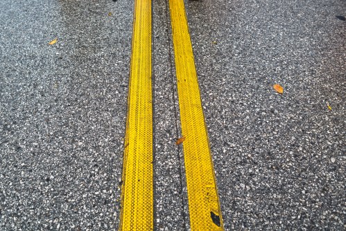 Two parallel yellow lines on a textured gray asphalt surface, with a few scattered leaves nearby.