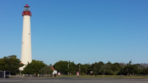 A tall lighthouse with a red top stands against a clear blue sky, surrounded by greenery and an empty parking lot.