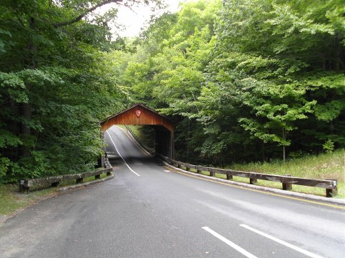 A scenic view of a covered bridge surrounded by lush green trees, leading to a winding road.