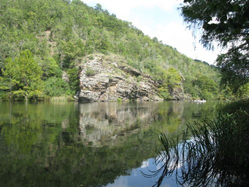 A serene river scene with a rocky cliff and lush greenery reflected in the calm water under a blue sky.